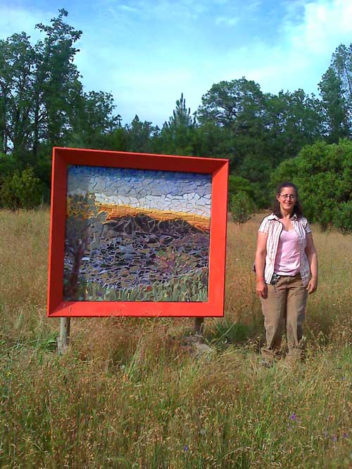 4 ft by 4 ft colored mosaic of a sunset view of Clearlake and Mt Konocti . The Artist Alicia Farnsworth stands beside it in a rural park setting.